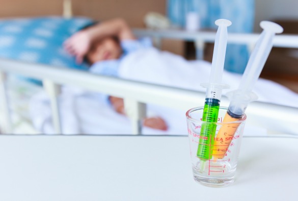 Syringes in a glass measuring cup with blurred illness boy lying on sickbed in hospital. Shallow depth of field (DOF), syringe in focus, child out of focus.
