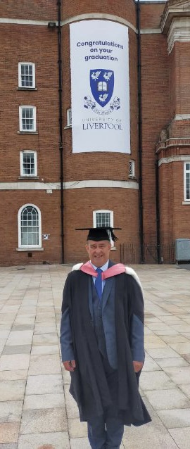 Phil in graduation robes with a university of Liverpool sign in the background