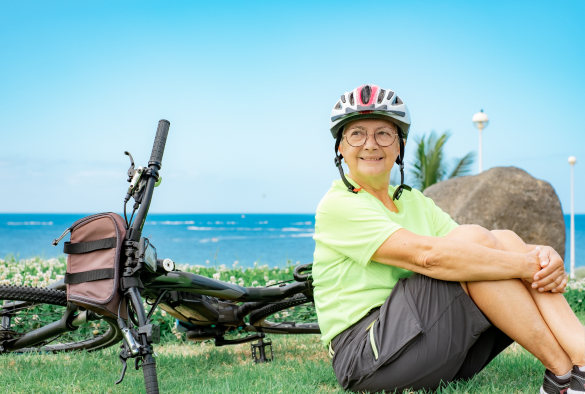 A woman in cycle helmet rests next to her bike, sea in background