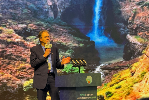Larry presenting at the work archaeological forum in Shanghai he is speaking into a microphone againsta colourful backdrop of a waterfall