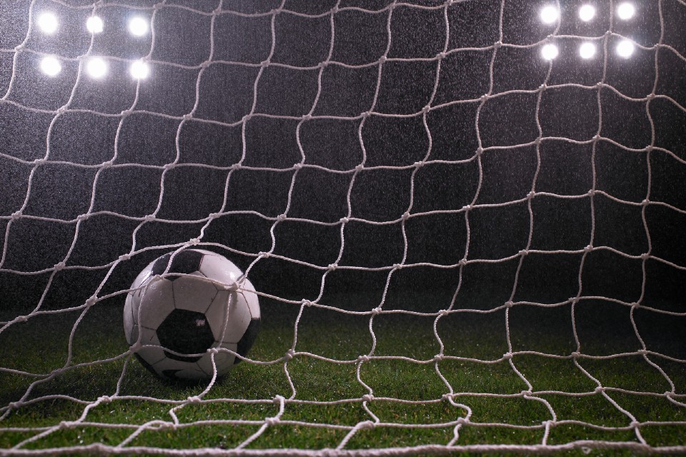 Football in goal net on floodlit pitch