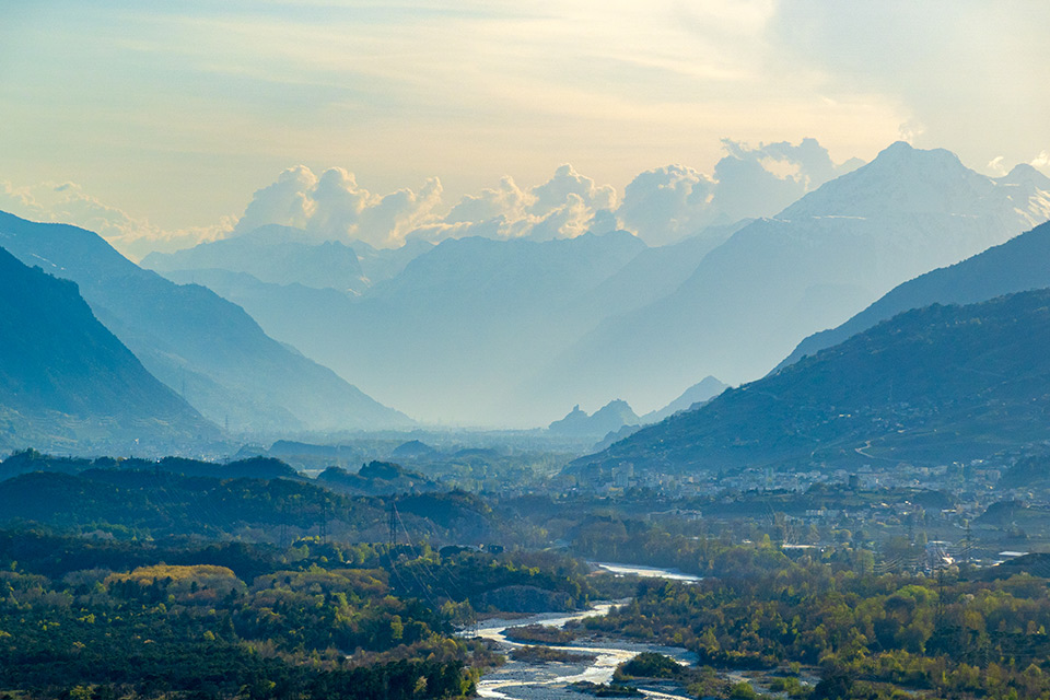 Scenery of a river winding through a green forest, with towering blue Swiss Alps on a cloudy day