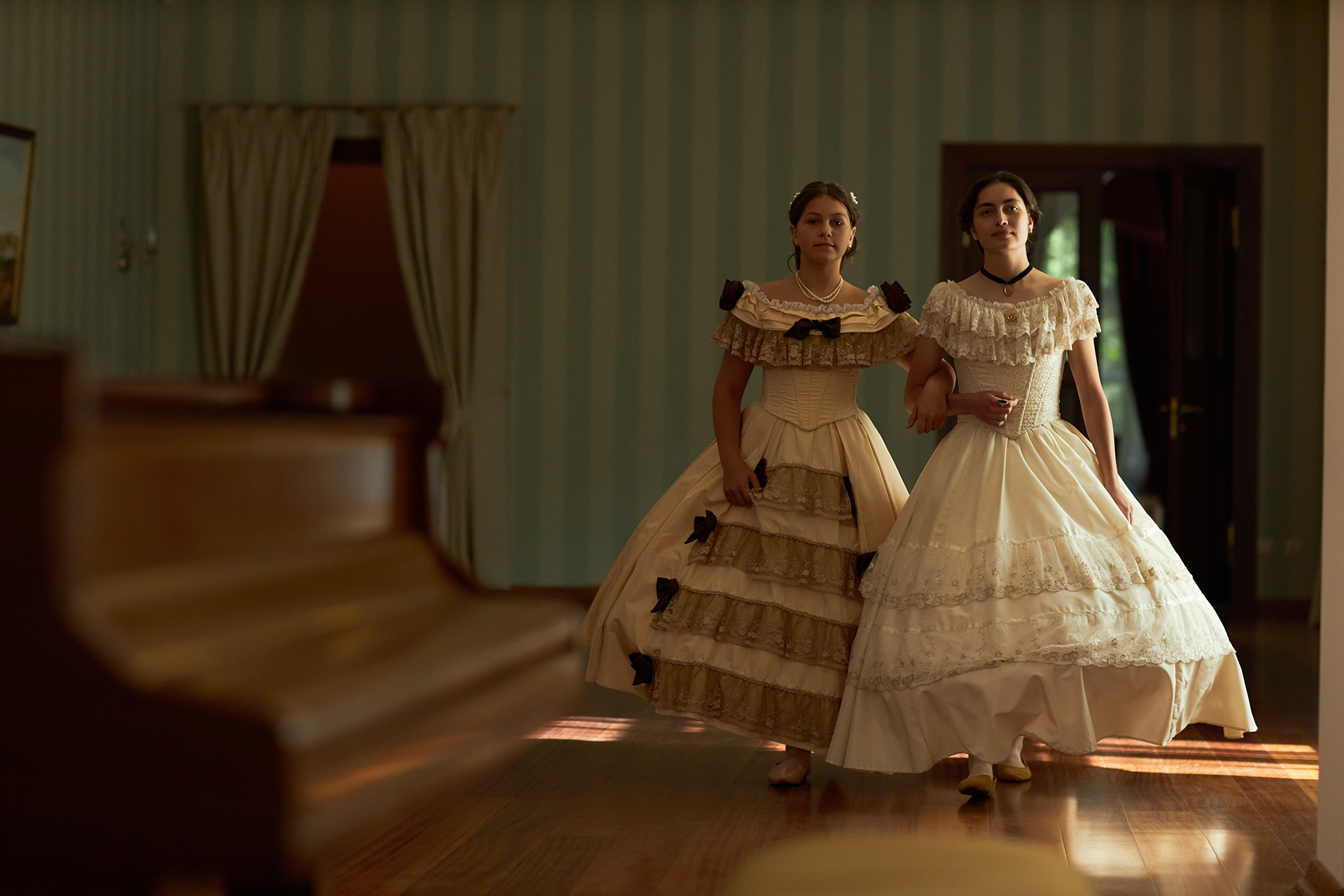 Full length portrait of two young ladies walking together towards camera in voluminous classic dresses across wooden floor in softly lit vintage ballroom