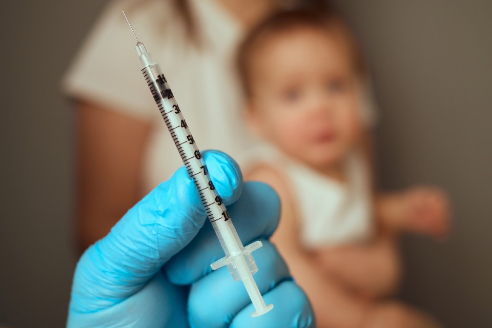 Stock image - gloved health worker's hand holding syringe, baby and parent/guardian in background